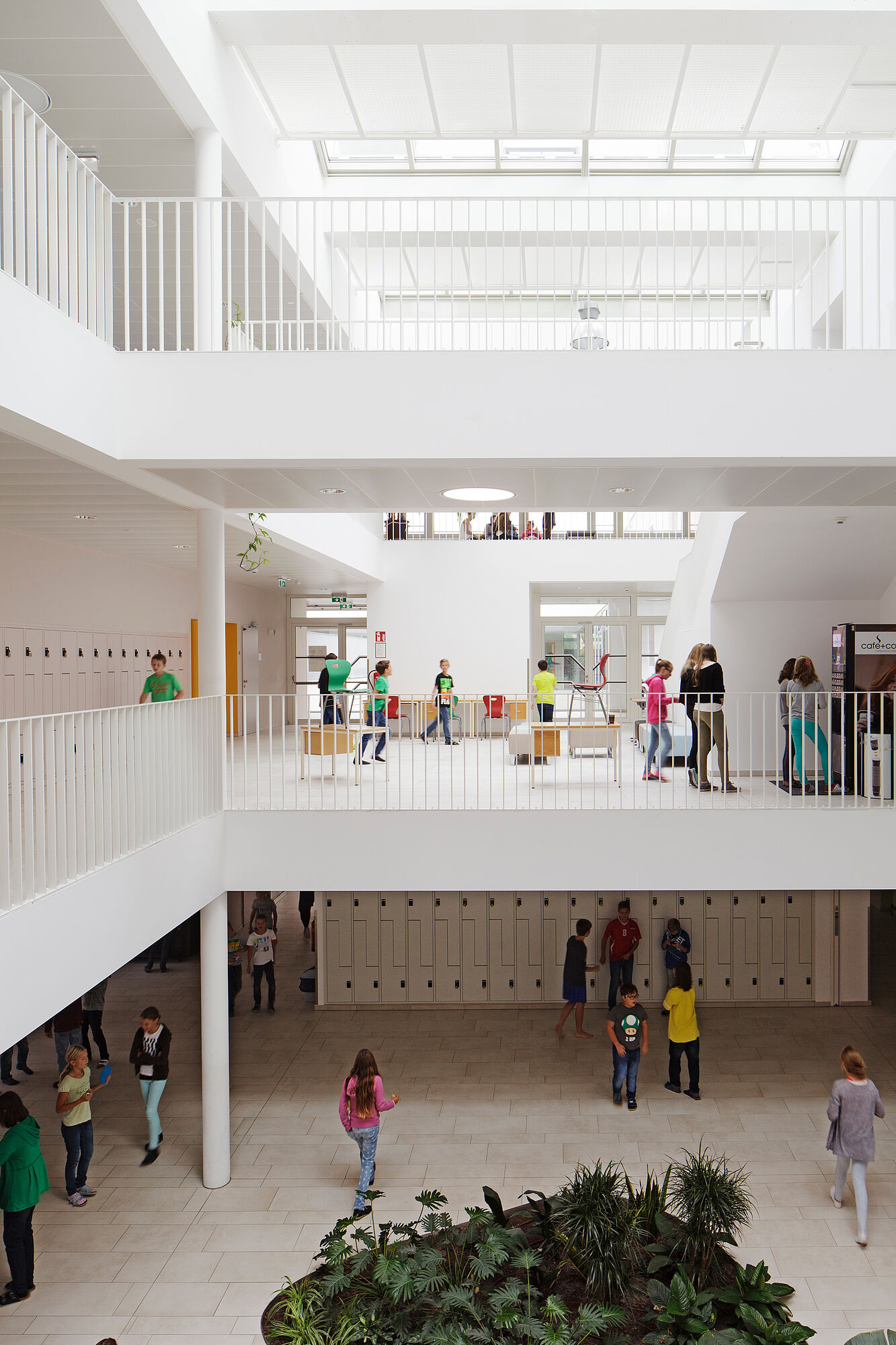 Three-story atrium lit from above, dominated by white; the students’ bright clothing adds colorful accents.